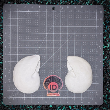 Two white resin plastic nautilus shells placed on a gray gridded cutting mat, with a small pink and black ID fabrications logo centered between them on a dark speckled surface.