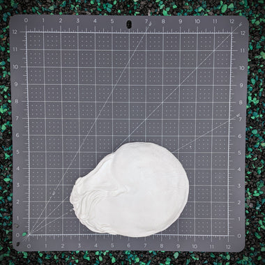 A white, plastic nautilus shell for mermaid tops placed on a gray self-healing cutting mat with grid lines and measurement markings, set against a background of black and green gravel.