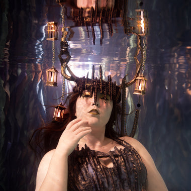 A woman underwater wearing a dark, skeletal or reptile-like costume top with dripping details and a matching headpiece with sharp protrusions, surrounded by hanging lanterns in a blue-toned underwater setting part of ID Fabrications Stillwater Collection