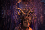 A person wearing an elaborate horned headpiece made of metallic spikes and shell-like elements, photographed underwater against a purple and blue marbled background from the stillwater collection