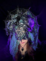 Person wearing an elaborate headpiece with spiderweb design, crab and purple accents on a dark background
