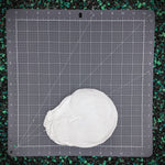 A white, plastic nautilus shell for mermaid tops placed on a gray self-healing cutting mat with grid lines and measurement markings, set against a background of black and green gravel.