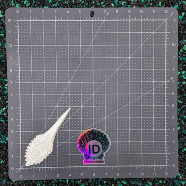 Gray cutting mat with grid lines, a white leaf-shaped object, and a colorful 'ID Fabrics' logo on a dark textured background.