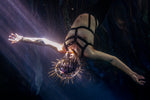 A person underwater wearing a black strappy harness-style bodysuit and a spiked crown-like headpiece, with dramatic light rays shining into the dark blue water from the stillwater collection