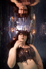 A woman underwater wearing a dark, skeletal or reptile-like costume top with dripping details and a matching headpiece with sharp protrusions, surrounded by hanging lanterns in a blue-toned underwater setting part of ID Fabrications Stillwater Collection