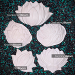 Four white sculpted lotus flower mermaid top DIY pieces arranged on a dark textured background with green and black pebbles. Each flower has a text label overlay: 'AwakenedBloom', 'GatheringBloom', 'KindredBloom', 'FirstBloom', and 'MorningBloom'.