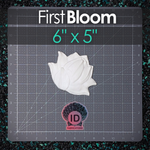 A white hand sculpted resin plastic lotus flower object displayed on a dark gray gridded cutting mat. The image features a dark overlay at the top with the text 'First Bloom' and '6" x 5"' in teal, and a logo for 'ID FABRICATIONS' near the bottom.