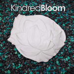 A white plastic resin flower with detailed petal textures for mermaid tops, displayed on a bed of black and green decorative gravel. The brand name 'KindredBloom' is written in white text at the top.