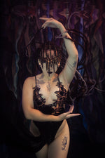 A model wearing a black lace and metallic gothic bodysuit with a matching headpiece, posed against a dark, abstract, purple-toned background part of the ID fabrications stillwater collection