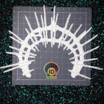 A white hand cast plastic skeletal or spiky decorative object arranged in a semi-circular shape on a gray gridded cutting mat, with a visible ID Fabrications logo near the bottom center. The mat is placed on a dark speckled surface.