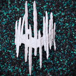 A white textured, spiky decorative base for a blind mask crown resembling icicles or a stylized crown, photographed from above on a black and teal pebble surface.