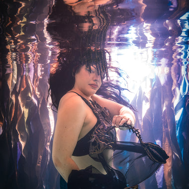 A woman underwater wearing a black and gold patterned silicone mermaid top and sheer black skirt, holding a black bonefish fan. The water creates strong reflections and ripples around her by ID Fabrications.