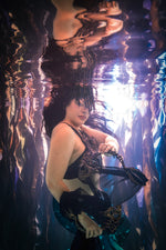 A woman underwater wearing a black and gold patterned silicone mermaid top and sheer black skirt, holding a black bonefish fan. The water creates strong reflections and ripples around her by ID Fabrications.